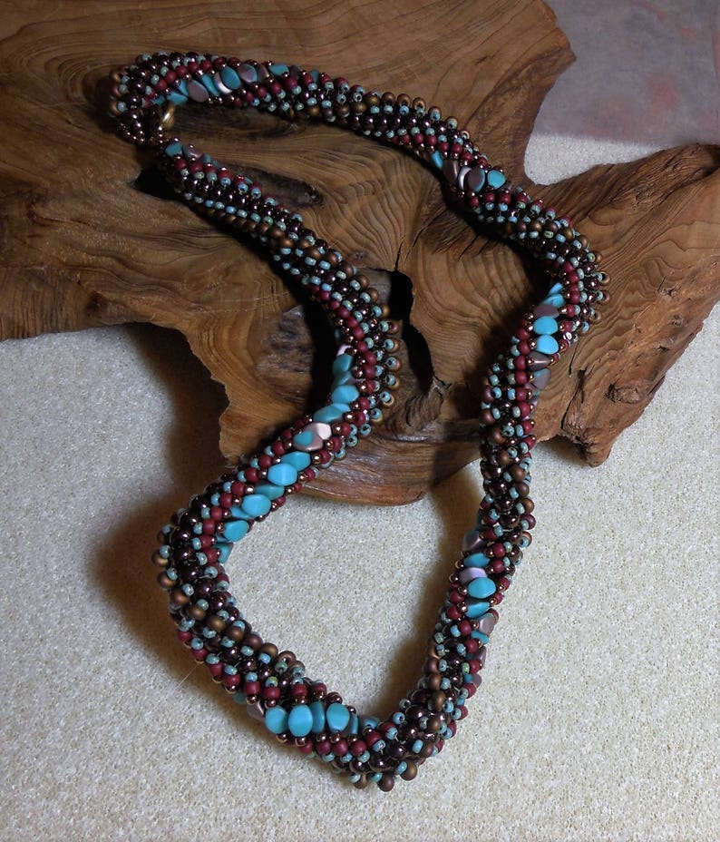 May include: A brown, red, and turquoise beaded necklace with a twisted design. The necklace is made of small, round beads and is displayed on a wooden surface.