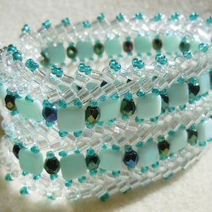 May include: A beaded bracelet with a woven design featuring alternating rows of clear and turquoise beads. The bracelet has a delicate and intricate look.