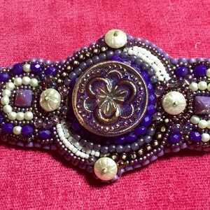 May include: A beaded bracelet with a purple and white design. The bracelet features a large, round, purple button with a floral design in the center.