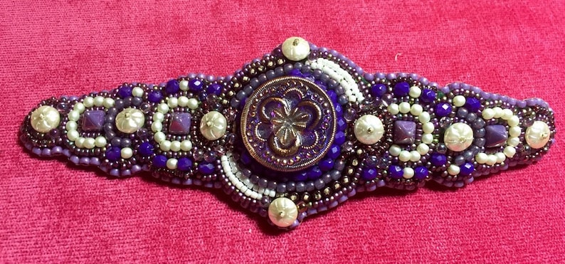 May include: A beaded bracelet with a purple and white design. The bracelet features a large, round, purple button with a floral design in the center.