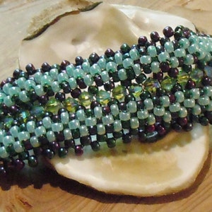 May include: A multi-strand beaded bracelet with a woven pattern in shades of teal, green, and purple. The bracelet is made with small seed beads and features a subtle shimmer.