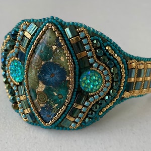 May include: A teal and gold beaded bracelet with a large, green, blue, and gold floral design in the center. The bracelet is made with a variety of beads, including seed beads, bugle beads, and crystals.