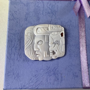 May include: A white carved pendant featuring two faces with intricate details. The pendant is made of a smooth, polished material and has a slightly off-white color.