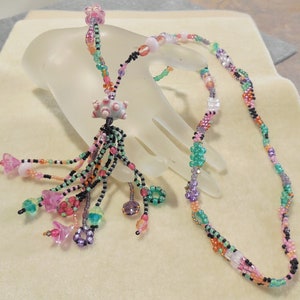 Puede incluir: Un collar de cuentas multicolores con un borla. El collar está hecho con cuentas pequeñas en tonos de rosa, verde, morado y naranja. La borla está hecha con las mismas cuentas y tiene una cuenta grande de color rosa en forma de estrella en la parte superior.