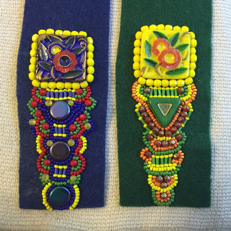 May include: Two beaded embellishments with colorful beads and a ceramic tile with a floral design. The embellishments are on a blue and green fabric background.