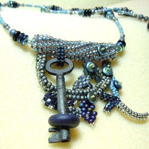 Puede incluir: Un collar de cuentas plateadas y azules con un gran colgante de llave antigua. La llave es de metal y tiene una cuenta de vidrio azul oscuro en el extremo del mango.