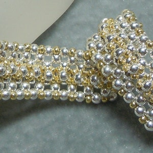 Puede incluir: Un brazalete de cuentas plateadas y doradas con un patrón tejido. El brazalete está hecho con cuentas pequeñas y redondas y tiene un cierre de broche.