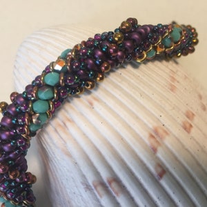 May include: A close-up of a beaded bracelet with purple, gold, and turquoise beads. The bracelet is on a white, textured surface.