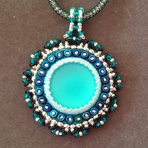 May include: A turquoise blue glass pendant with a beaded border in shades of teal, black, and silver. The pendant is attached to a black mesh chain.