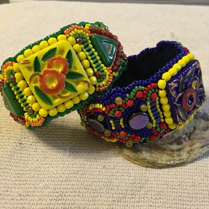 May include: Two beaded bracelets with colorful geometric patterns and floral designs. The bracelets are made with yellow, blue, green, red, and orange beads.