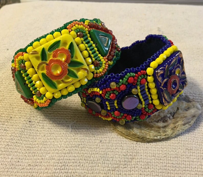 May include: Two beaded bracelets with colorful geometric patterns and floral designs. The bracelets are made with yellow, blue, green, red, and orange beads.
