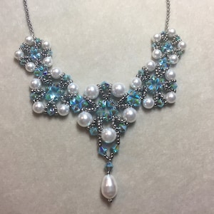 May include: A silver chain necklace with a teardrop-shaped pendant made of white pearls and blue iridescent crystals. The necklace features a geometric design with multiple layers of crystals and pearls.