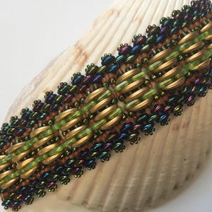 May include: A beaded bracelet with a woven pattern of gold, green, and iridescent beads. The bracelet is displayed on a seashell.