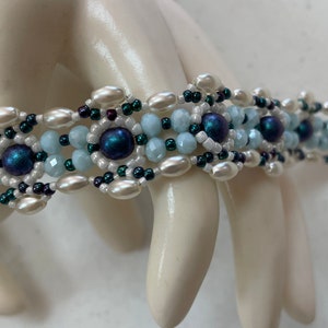May include: A beaded bracelet featuring a repeating pattern of white, light blue, and dark blue beads. The bracelet is made with a variety of bead shapes, including round and oval, and has a silver clasp. The bracelet is approximately 7 inches long.