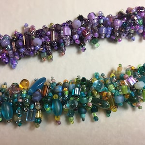 May include: Two strands of beaded jewelry with a variety of colors and shapes. The top strand is purple, lavender, and silver. The bottom strand is green, blue, and gold.