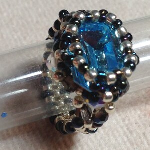 May include: A beaded ring featuring a large, rectangular, light blue gemstone. The ring is surrounded by a beaded border of silver, black, and iridescent beads. The ring is displayed on a clear, cylindrical object.