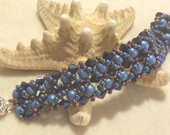 May include: A beaded bracelet featuring a woven pattern of dark blue and black beads, accented with small metallic brown beads. The bracelet includes a rectangular, clear crystal-encrusted clasp with a gold-toned metal setting at one end.
