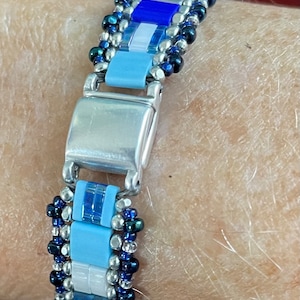 May include: A blue, white, and silver beaded bracelet with a silver clasp. The bracelet is made with small, square beads and has a geometric pattern.