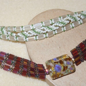 Puede incluir: Dos pulseras de cuentas, una con cuentas blancas y verdes y la otra con cuentas rojas y marrones. La pulsera roja y marrón tiene un gran colgante de vidrio de colores.