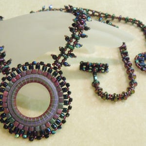 May include: A handcrafted beaded necklace set with a large circular pendant. The necklace, clasp, and pendant are decorated with purple, green, and black beads. The necklace is a unique piece of jewellery.