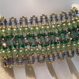 May include: A beaded bracelet with green, gold, and clear beads. The bracelet is rectangular, with rows of round green beads, rectangular green beads, and gold accents. Clear beads are rectangular and round. The bracelet is approximately 18 cm in length.