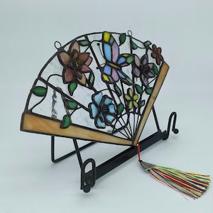 May include: Stained glass fan with a butterfly and floral design in shades of pink, blue, yellow, and green. The fan is displayed on a black metal stand and has a colorful tassel. The fan is a decorative home decor item.