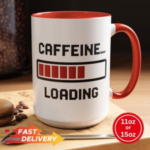 May include: White ceramic coffee mug with a red interior and handle. The mug features the text "CAFFEINE... LOADING" with a loading bar graphic. A red sticker indicates the mug is available in 11oz or 15oz sizes.