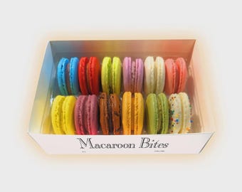 May include: A box of colorful macarons, including blue, red, yellow, green, and pink. The macarons are arranged in rows inside a white box with the text "Macaroon Bites" printed on the front.