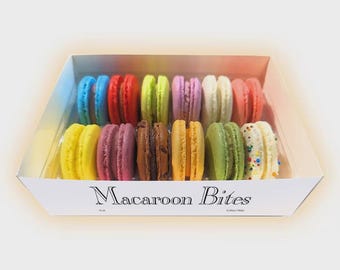 May include: A box of colorful macarons, including blue, red, yellow, green, purple, and brown. The box is white with the words "Macaroon Bites" printed on the front. The macarons are arranged in rows.