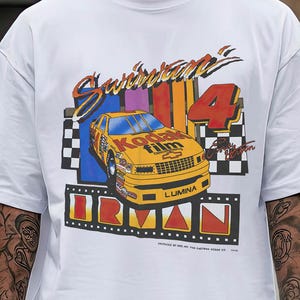 May include: White t-shirt featuring a colorful graphic of a yellow race car with the text "Swanson 4" and "Irvan Lumina". The design includes a checkered flag and the Kodak Film logo. The shirt has short sleeves.