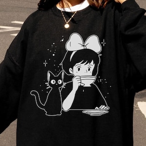 以下が含まれることがあります： 黒いスウェットシャツ。白いグラフィックは、リボンをした女の子、黒猫、ティーカップ。スウェットシャツはクルーネックと長袖です。「FAST DELIVERY」の文字が左上に赤と白で表示されています。