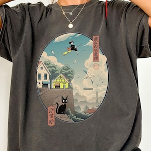 May include: Pepper-colored t-shirt featuring a circular graphic with Japanese art. The graphic depicts a witch flying on a broom, a black cat, and traditional Japanese buildings. The text on the shirt is in Japanese.