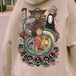 May include: Beige hoodie with a Japanese animated film graphic. The design features a girl with a lantern, a dragon, and other characters. The hoodie has a drawstring hood and long sleeves.