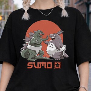 Könnte beinhalten: Schwarzes T-Shirt mit einem Aufdruck von Godzilla und Totoro beim Sumo-Ringen vor einem roten Kreis. Das Wort "SUMO" ist darunter gedruckt. Das T-Shirt hat eine lockere, übergroße Passform.