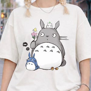 May include: Ivory-colored, oversized t-shirt featuring a graphic of a gray Totoro character holding a flower, with a smaller blue Totoro. The shirt has short sleeves and the text "IVORY COMFORT COLORS".