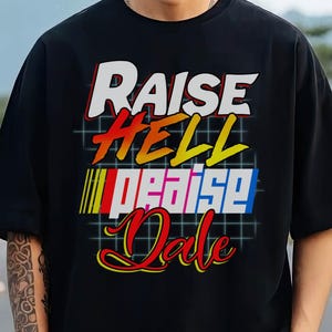 May include: Black t-shirt with the text "RAISE HELL PRAISE Dale" in a retro font. The text is in white, red, yellow, and blue. The shirt has short sleeves and a crew neck.