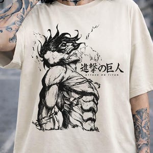 May include: Cream-colored t-shirt featuring a black and white illustration of a muscular figure with the text "Attack on Titan" in Japanese and English. The graphic design is centered on the front of the shirt.