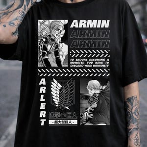 Op de afbeelding: Zwart T-shirt met anime-achtige graphics en tekst. Het ontwerp bevat de naam "ARMIN" herhaald, samen met de tekst "TO ENDURE BECOMING A MONSTER YOU HAVE TO DISCARD YOUR HUMANITY." Bevat ook de tekst "ARLERT" en Japanse tekst.