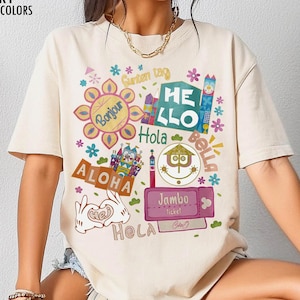 May include: Ivory-colored t-shirt with a colorful graphic design featuring the words "Hello", "Aloha", "Bonjour", and "Hola". The design includes floral and cartoon elements, and the text "Jambo Ticket".