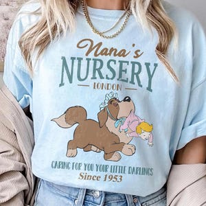 Pode incluir: Camiseta azul clara com o texto "Nana's Nursery London" e uma ilustração de desenho animado de um cachorro segurando um bebê. A camisa também inclui o texto "Caring for you your little darlings Since 1953".