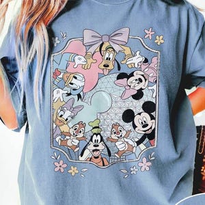 Könnte beinhalten: Blaues Jeans-T-Shirt mit einem farbenfrohen Aufdruck von Disney-Figuren wie Mickey Mouse, Minnie Mouse, Donald Duck und Goofy. Die Grafik ist mit einer rosa Schleife und floralen Akzenten umrahmt.