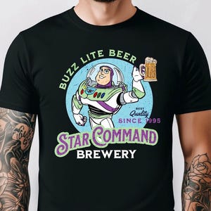 Op de afbeelding: Zwart t-shirt met een cartoon illustratie van Buzz Lightyear die een bierpul vasthoudt. Het ontwerp bevat de tekst "Buzz Lite Beer" en "Star Command Brewery" met "Best Quality Since 1995".