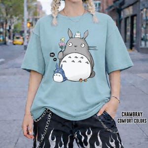 May include: A light blue t-shirt featuring a graphic of Totoro and other characters from the animated film My Neighbor Totoro. The design includes a large gray Totoro holding a flower, a small blue Totoro, and other small black creatures. The text "CHAMBRAY COMFORT COLORS" is printed on the shirt.