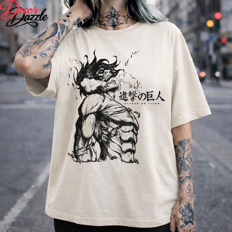 May include: Cream-colored t-shirt featuring a black and white illustration of a muscular figure with the text "Attack on Titan" in Japanese and English. The graphic design is centered on the front of the shirt.