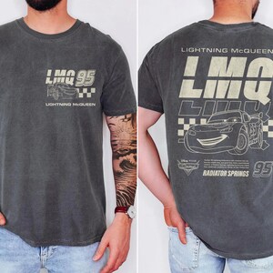 Puede incluir: Camiseta gris oscuro con Lightning McQueen de la pel&iacute;cula Cars. La parte delantera muestra "LMO 95" con un gr&aacute;fico de coche y "Lightning McQueen". La parte trasera tiene un dise&ntilde;o m&aacute;s grande con el coche, "95" y "Radiator Springs."