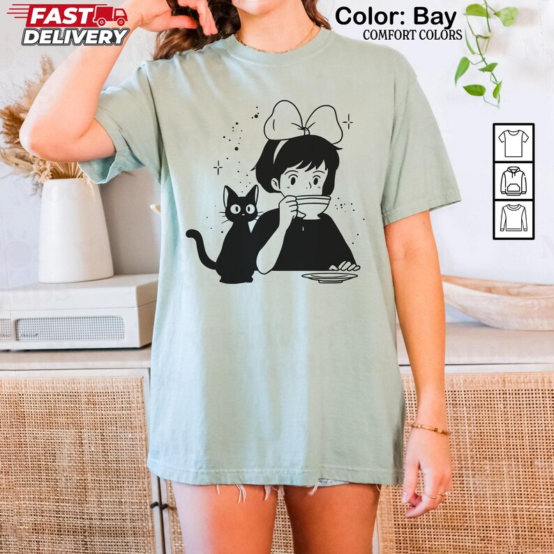 以下が含まれることがあります： 女の子と黒猫のイラストがプリントされた、薄緑色のTシャツ。女の子はリボンをつけ、カップから飲み物を飲んでいます。「Color: Bay」と「Comfort Colors」の文字が上部にプリントされています。半袖です。