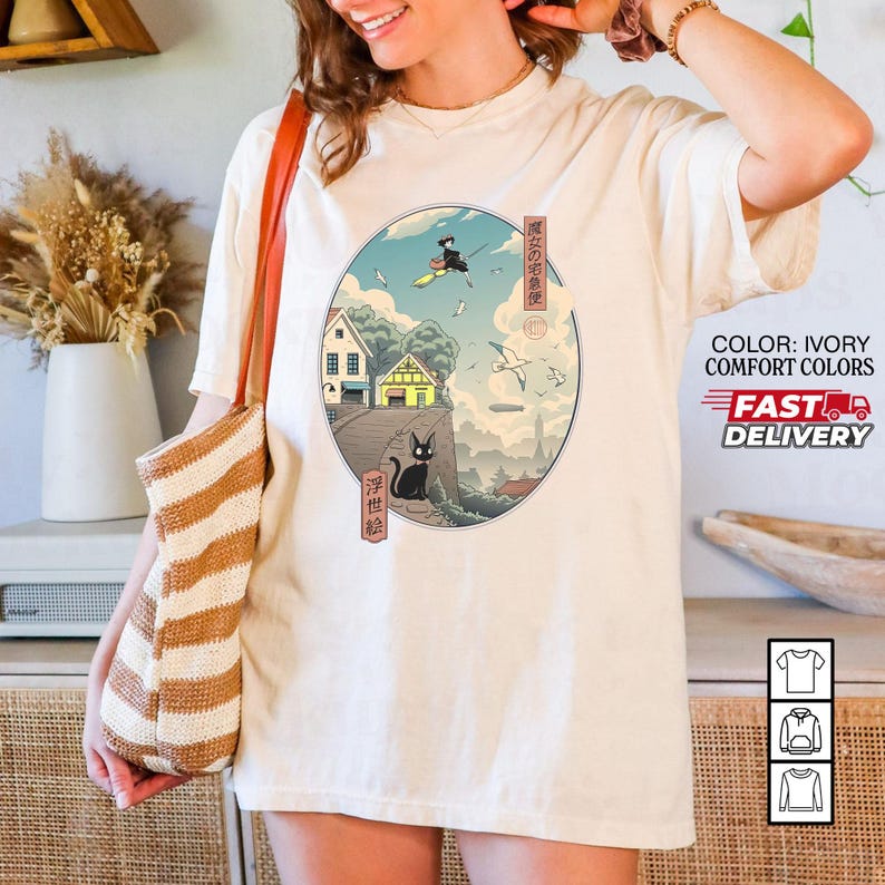 May include: Ivory-colored t-shirt featuring a circular graphic with a Japanese-inspired illustration. The design includes a witch on a broom, a black cat, houses, and text in Japanese. The shirt also has the text "COLOR: IVORY COMFORT COLORS" and "FAST DELIVERY".