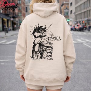 May include: A beige hoodie with a black and white graphic of a muscular figure from the anime series "Attack on Titan." The Japanese text is also visible. The hoodie has a hood and long sleeves.