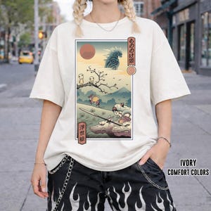 May include: Ivory-colored t-shirt featuring a Japanese anime-style illustration with a red sun, mountains, and characters. The graphic includes Japanese text and is framed within a rectangular border. The shirt has short sleeves.