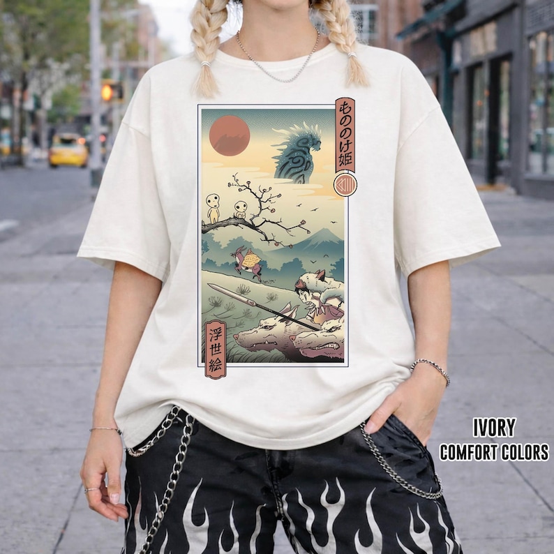 May include: Ivory-colored t-shirt featuring a Japanese anime-style illustration with a red sun, mountains, and characters. The graphic includes Japanese text and is framed within a rectangular border. The shirt has short sleeves.